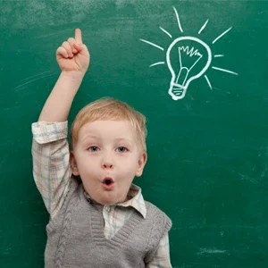 A young child holding his hand up to answer a question. A lightbulb is drawn on the chalkboard behind him